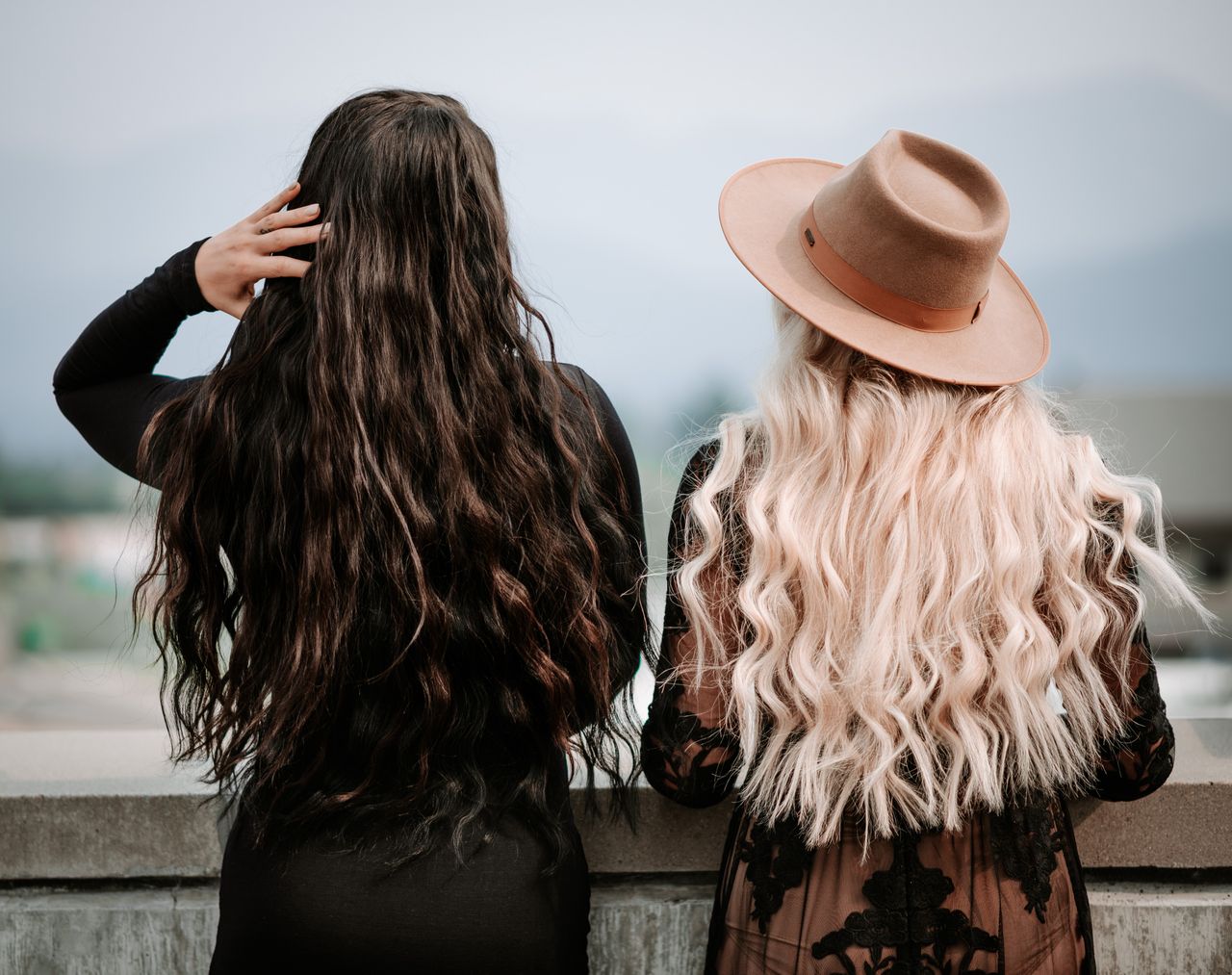 Two stylish women with long wavy hair standing on a rooftop, one in a fedora.
